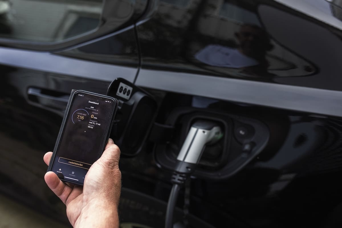 En hånd holder opp en mobiltelefon og viser frem et bilde fra en app. I bakgrunnen en elbil som lader.