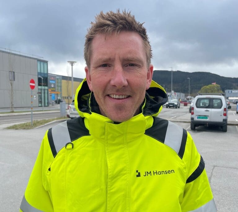 Smilende elektriker med JM Hansen.logoen på jakken stor foran en parkeringsplass og et næringsbygg