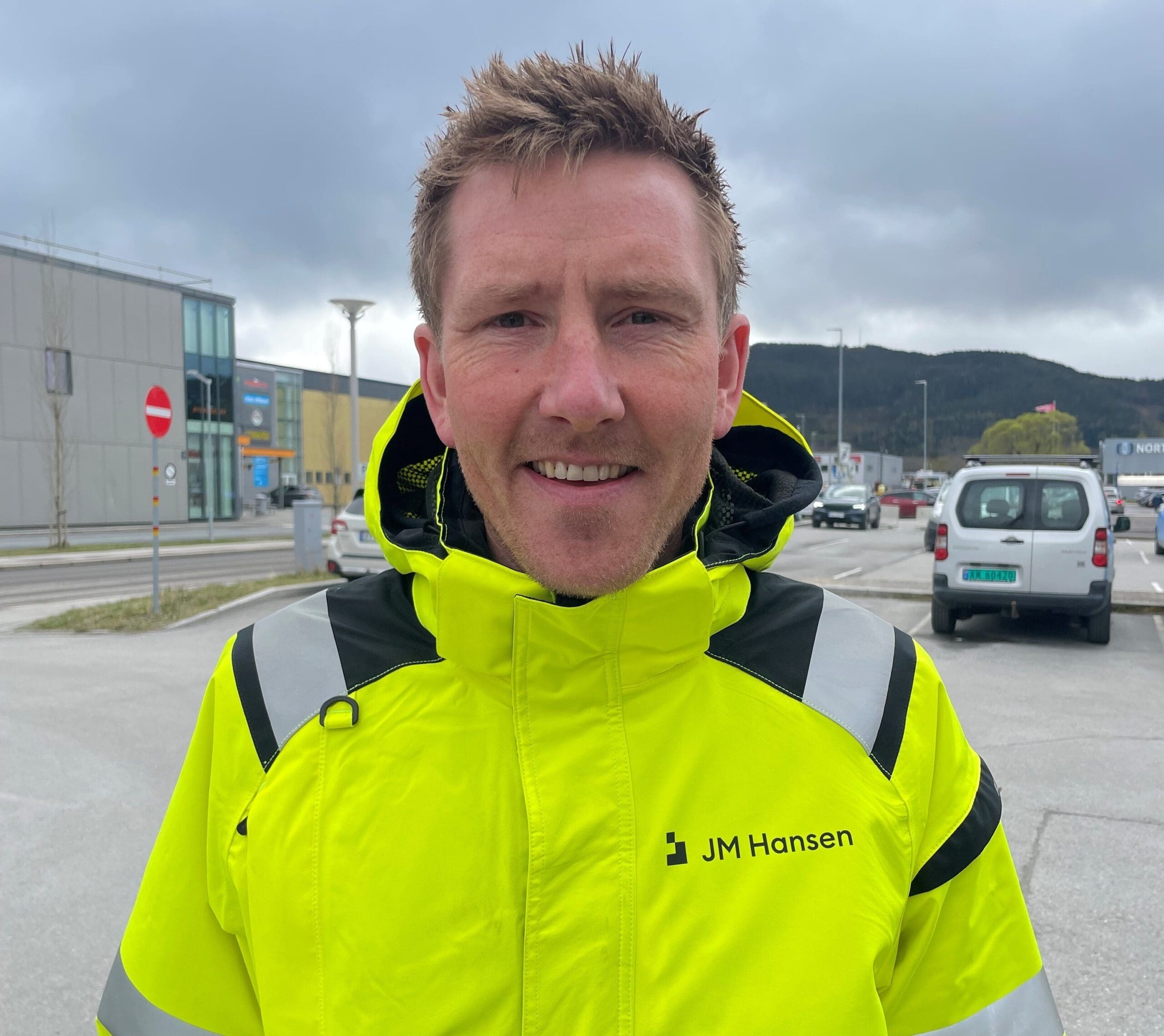 Smilende elektriker med JM Hansen.logoen på jakken stor foran en parkeringsplass og et næringsbygg